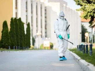 Pest control professional in full protective white Hazmat suit and mask spraying pesticide along an outdoor walkway.