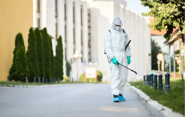 Pest control professional in full protective white Hazmat suit and mask spraying pesticide along an outdoor walkway.