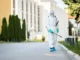 Pest control professional in full protective white Hazmat suit and mask spraying pesticide along an outdoor walkway.