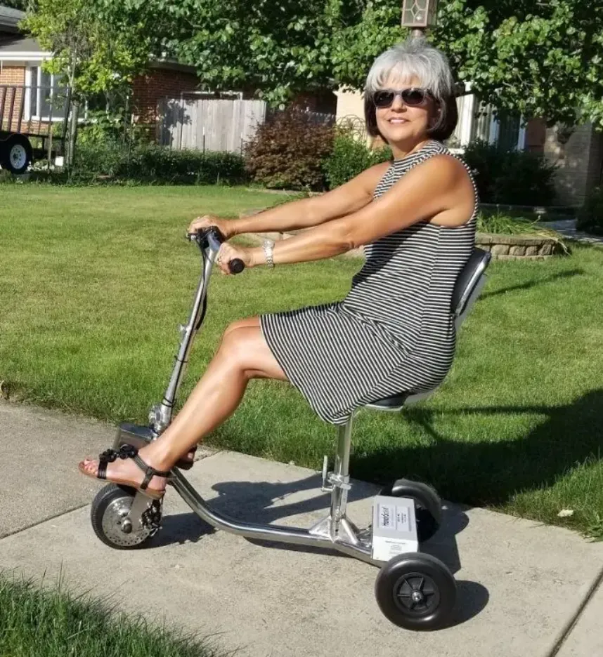 Senior woman wearing sunglasses riding a lightweight, compact 3-wheel folding mobility scooter on a sunny sidewalk.