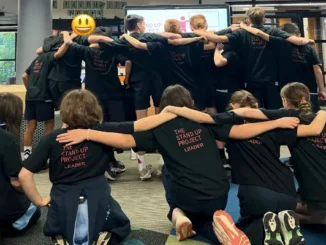 A group of students wearing matching black t-shirts that read "The Stand Up Project Leader" standing and kneeling in a circle with their arms around each other's shoulders during a leadership training workshop.