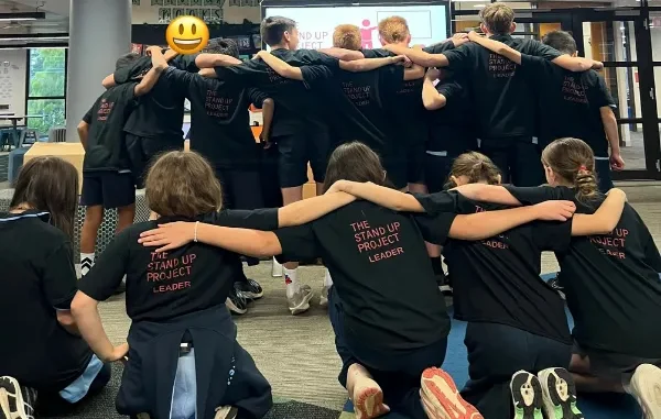 A group of students wearing matching black t-shirts that read "The Stand Up Project Leader" standing and kneeling in a circle with their arms around each other's shoulders during a leadership training workshop.