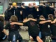 A group of students wearing matching black t-shirts that read "The Stand Up Project Leader" standing and kneeling in a circle with their arms around each other's shoulders during a leadership training workshop.