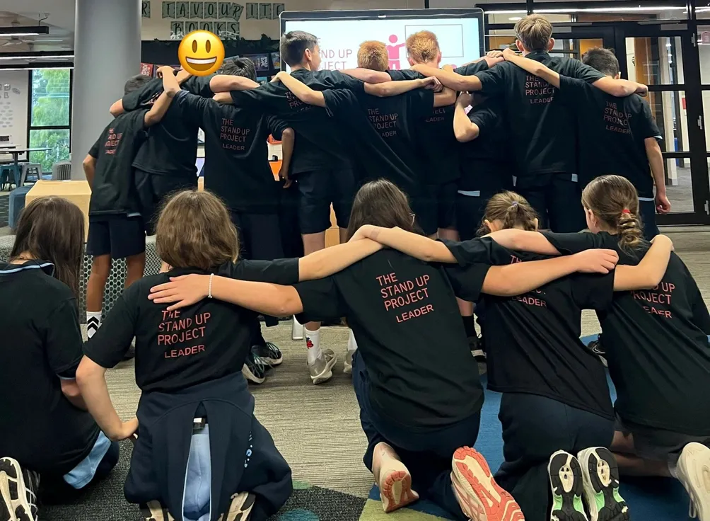 Group of students kneeling and standing in a supportive huddle wearing matching 'The Stand Up Project Leader' t-shirts during a student leadership training activity.