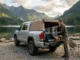 A silver Toyota Tacoma with a Softopper canvas camper shell installed, parked by a mountain lake at sunset. A man accesses the open tailgate, which is organized with camping gear and storage bins.