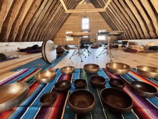 A group of people lying down on yoga mats during a sound healing meditation session in a wooden A-frame barn attic, with Tibetan singing bowls arranged on a colorful blanket in the foreground.