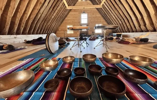A group of people lying down on yoga mats during a sound healing meditation session in a wooden A-frame barn attic, with Tibetan singing bowls arranged on a colorful blanket in the foreground.