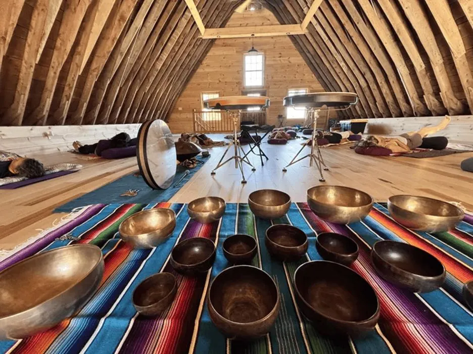 A group of people participating in a sound healing session during a wellness retreat, lying down on mats under a high wooden A-frame ceiling while various singing bowls and drums rest on a colorful blanket in the foreground.