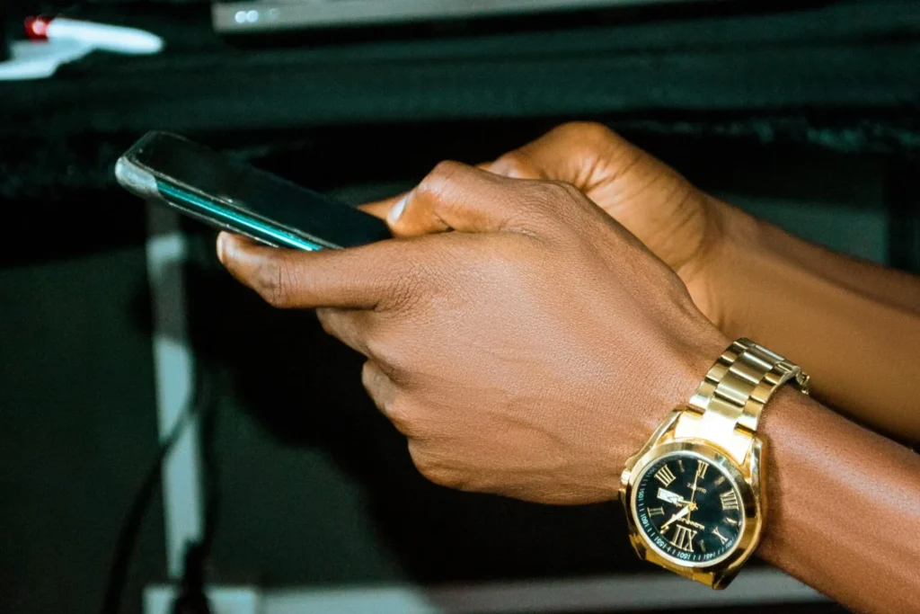 Close-up of a person's hands wearing a gold watch while using a smartphone to place an accumulator bet on a Zambian sports betting platform.