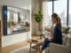 A woman sitting in a bright, modern living room using a digital tablet to interact with an AI 3D room design interface displayed on a large screen wall mount.