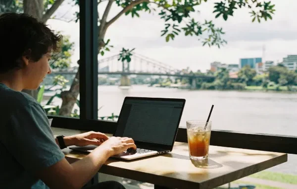 A digital nomad working on a laptop at a cafe table next to an iced drink, with the Brisbane River and Story Bridge visible outside the window.