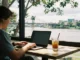 A digital nomad working on a laptop at a cafe table next to an iced drink, with the Brisbane River and Story Bridge visible outside the window.