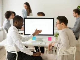 Two male professionals discussing a project on a computer screen in a bright modern office with colleagues working in the background.