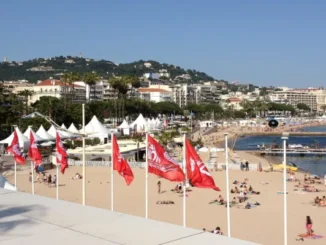 A high-angle view of a sandy beach in Cannes, France, featuring red festival flags flying in the foreground, people relaxing on the shore, and white event tents set up along the coast against a background of city buildings and green hills.