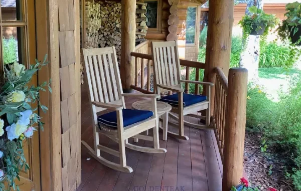 Two wooden teak rocking chairs with blue cushions placed on a rustic cabin porch with a small side table.
