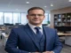 Professional headshot of Dr. Luiz Pacheco wearing glasses and a blue three-piece suit in a modern office environment.