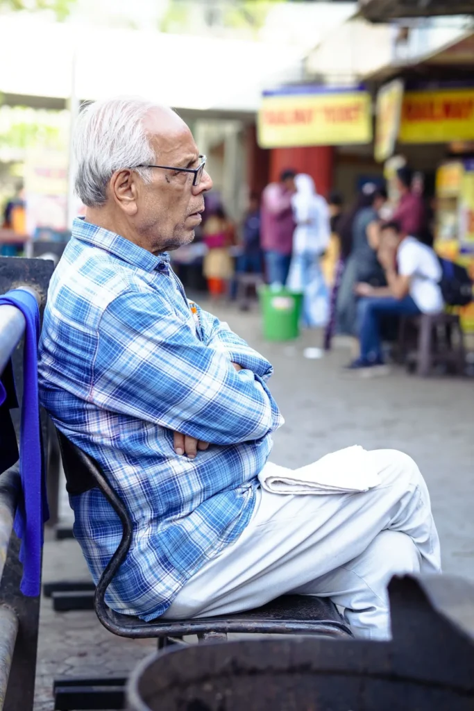 A medium side profile photograph of a pensive elderly man with white hair and glasses, wearing a blue and white checkered shirt and light pants, seated outdoors with his arms and legs crossed, looking to the side in a public space.
