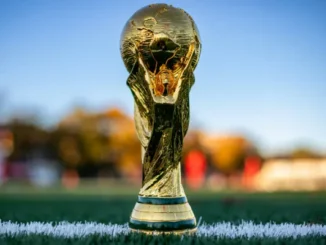 Close-up of the official golden FIFA World Cup trophy standing on the white touchline of a green soccer pitch.