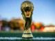 Close-up of the official golden FIFA World Cup trophy standing on the white touchline of a green soccer pitch.