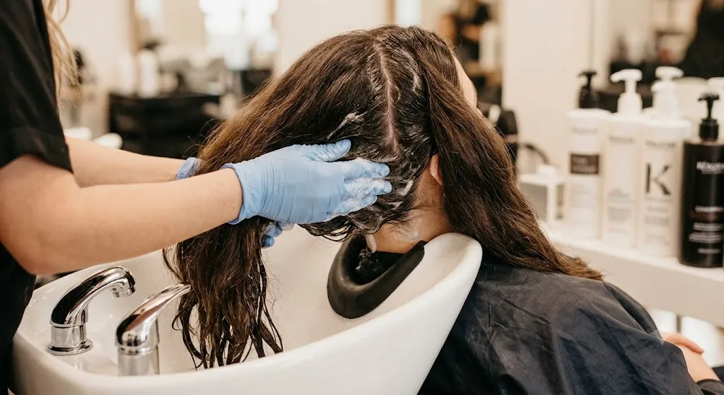 Professional hair stylist applying shampoo directly to a client's scalp at a salon wash basin while keeping thick hair strands out of the focus area.