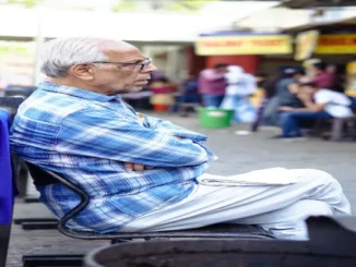 An elderly man with white hair and glasses wearing a blue plaid shirt, sitting outdoors with his arms crossed in a public setting.