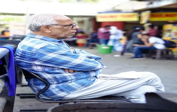 An elderly man with white hair and glasses wearing a blue plaid shirt, sitting outdoors with his arms crossed in a public setting.