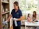 A uniformed Shorely Clean professional dusts a bookshelf while a mother and daughter relax and play a puzzle in a bright, clean living room.
