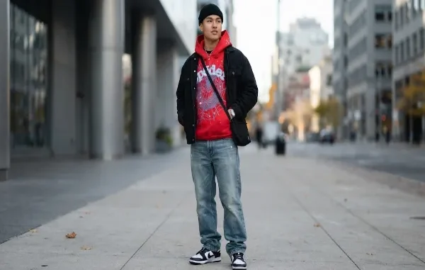 A young man modeling a streetwear outfit on a city sidewalk, wearing a vibrant red Sp5der Worldwide hoodie with signature web graphics, layered under a black denim jacket, paired with light wash loose-fit denim jeans, a black beanie, a crossbody bag, and clean sneakers.