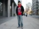 A young man modeling a streetwear outfit on a city sidewalk, wearing a vibrant red Sp5der Worldwide hoodie with signature web graphics, layered under a black denim jacket, paired with light wash loose-fit denim jeans, a black beanie, a crossbody bag, and clean sneakers.