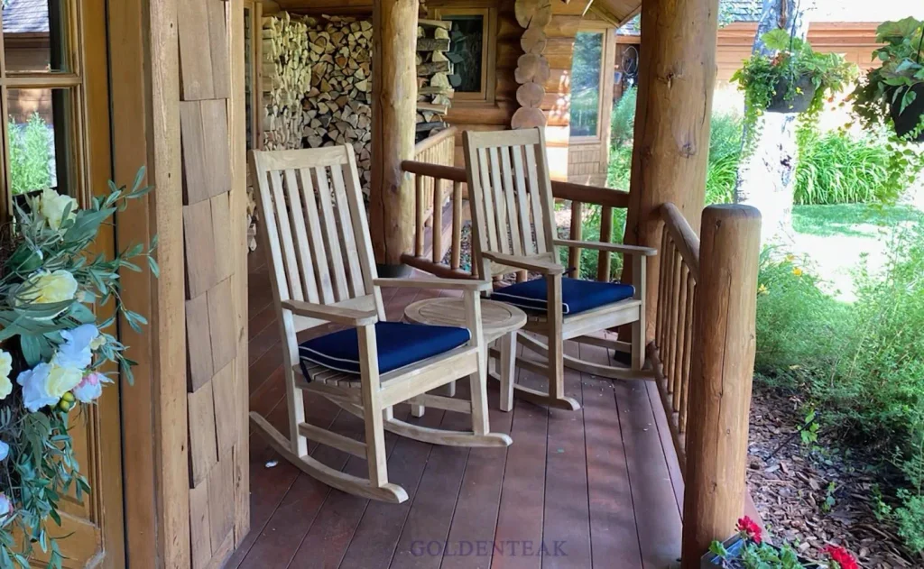 A pair of light-wood rocking chairs with navy blue seat pads sit on a rustic porch beside a small round table, with a stacked wood pile visible in the background.