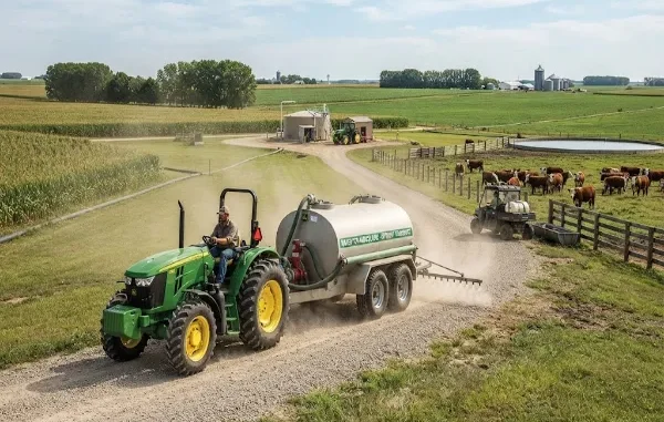 A green tractor pulling a large water tank trailer on a gravel farm road, with cattle and agricultural facilities visible in the background.
