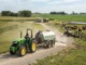 A green tractor pulling a large water tank trailer on a gravel farm road, with cattle and agricultural facilities visible in the background.