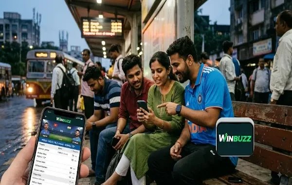 A close-up of a hand holding a smartphone displaying live cricket scores on the Winbuzz app, with a group of friends checking updates at a bus stop in the background.