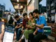 A close-up of a hand holding a smartphone displaying live cricket scores on the Winbuzz app, with a group of friends checking updates at a bus stop in the background.