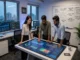 Three digital marketing professionals standing around an interactive touch-table analyzing colorful AI SEO analytics dashboards, with a whiteboard detailing AI-powered optimization strategies in the background.