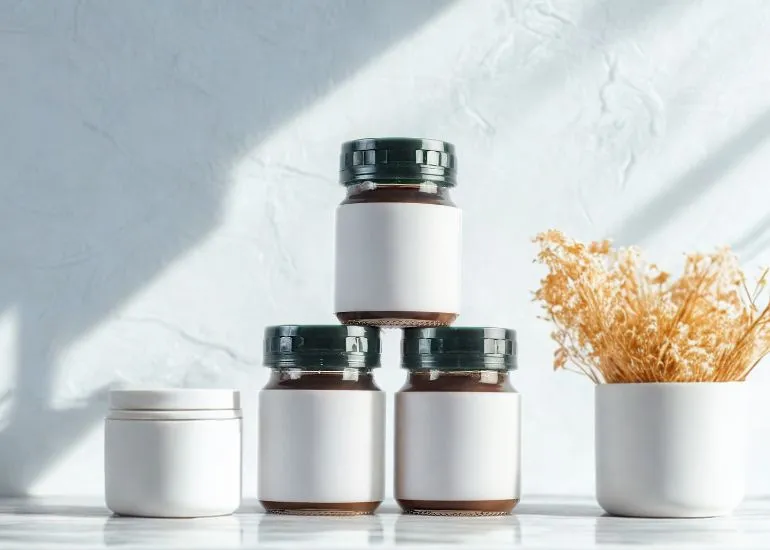 Three dark amber glass supplement bottles with prominent blank white labels are stacked in a pyramid. A white cosmetic jar is on the left, and a small vase with dried yellow flowers is on the right, all sitting on a marble table with sunny reflections and shadows on the textured white wall behind them.