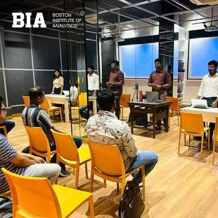 Students in orange chairs attend a presentation at the Boston Institute of Analytics (BIA) classroom, which features wooden floors, glass partitions, and a screen in a modern learning environment.