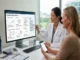 A female physician in a lab coat pointing at a Bioscope.ai Synoptic Health Dashboard on a desktop monitor while explaining personalized health data and longevity insights to a seated female patient.