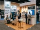 A well-lit, open-concept trade show booth featuring comfortable seating areas, clear signage, an interactive tablet demo, and friendly staff engaging with visitors.