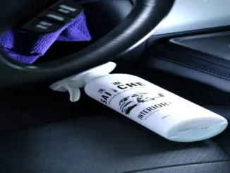 A white car interior cleaner spray bottle placed on a black leather seat next to a steering wheel and purple microfiber cloth.