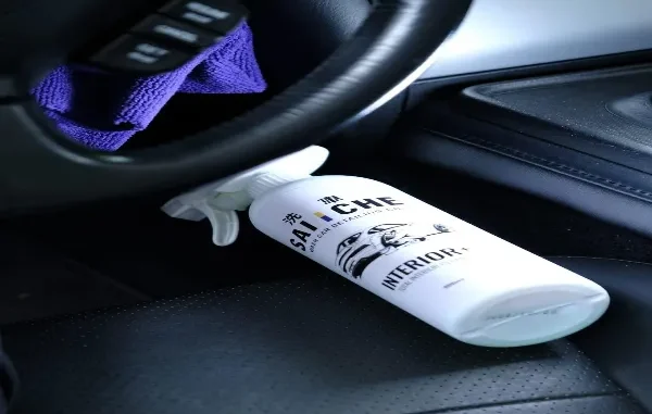 A white car interior cleaner spray bottle placed on a black leather seat next to a steering wheel and purple microfiber cloth.