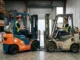 Side-by-side comparison in a warehouse of a shiny new orange forklift on the left and a dirty, heavily used old forklift on the right.