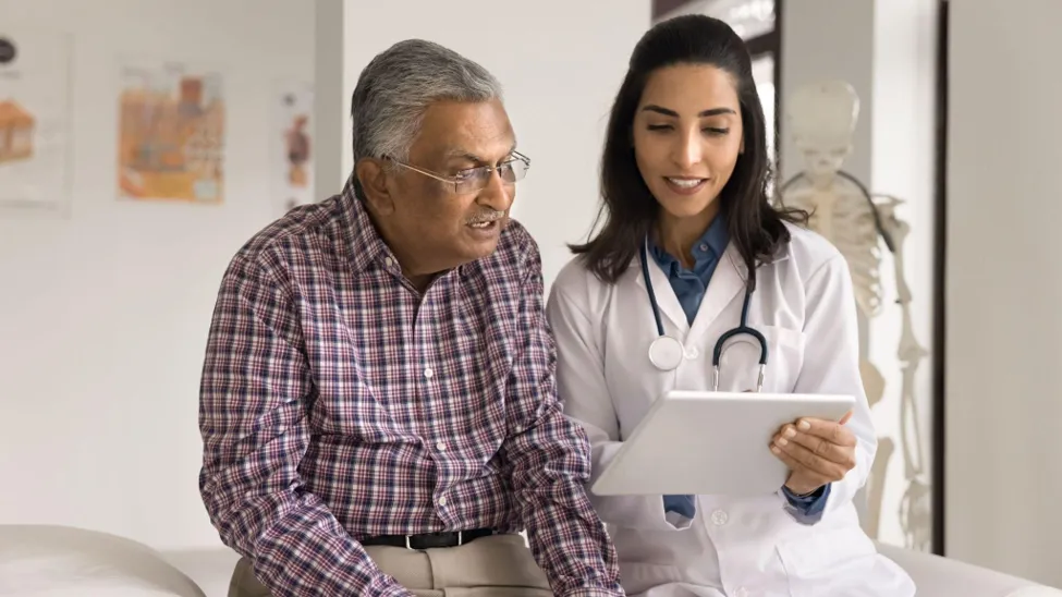 An elderly man in a plaid shirt and a female doctor in a white coat review a digital health plan on a tablet together in a clinic setting.