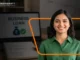Smiling Indian female entrepreneur standing in front of a laptop displaying a business loan approval graphic.