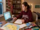 High school student sitting at a wooden desk researching colleges on a laptop with maps and brochures spread out.