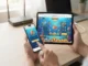 A close-up photograph of hands holding a smartphone and a tablet on a desk, both displaying the Fire Kirin online fish game directly within mobile web browsers, bypassing the need for app installation.