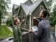 A homeowner in a green jacket points up at a roof valley where water is cascading over a gutter during a rainstorm. Beside her, a man wearing a jacket with the "Legacy Gutter Solutions" logo looks at the issue while holding a tablet showing a roof diagram. A company truck is on the right.