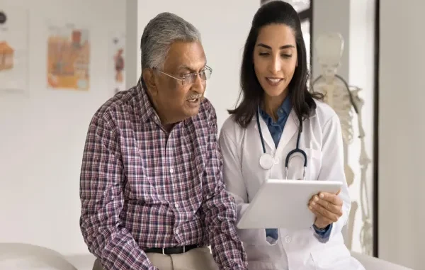 A smiling female doctor holding a tablet while explaining health insurance coverage options to an elderly man sitting in a clinic.