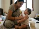 An Indian mother sitting on a bed, tenderly applying a specialized cream to her infant's arm, which shows visible signs of eczema and red spots.