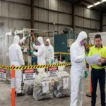 Industrial facility manager and contractor reviewing building schematics outside a safety-barricaded asbestos containment zone in a warehouse.